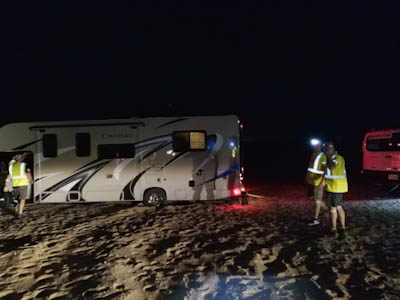 A white recreational vehicle is stuck in sand at night in Death Valley. Crew members ponder how to get the RV unstuck.
